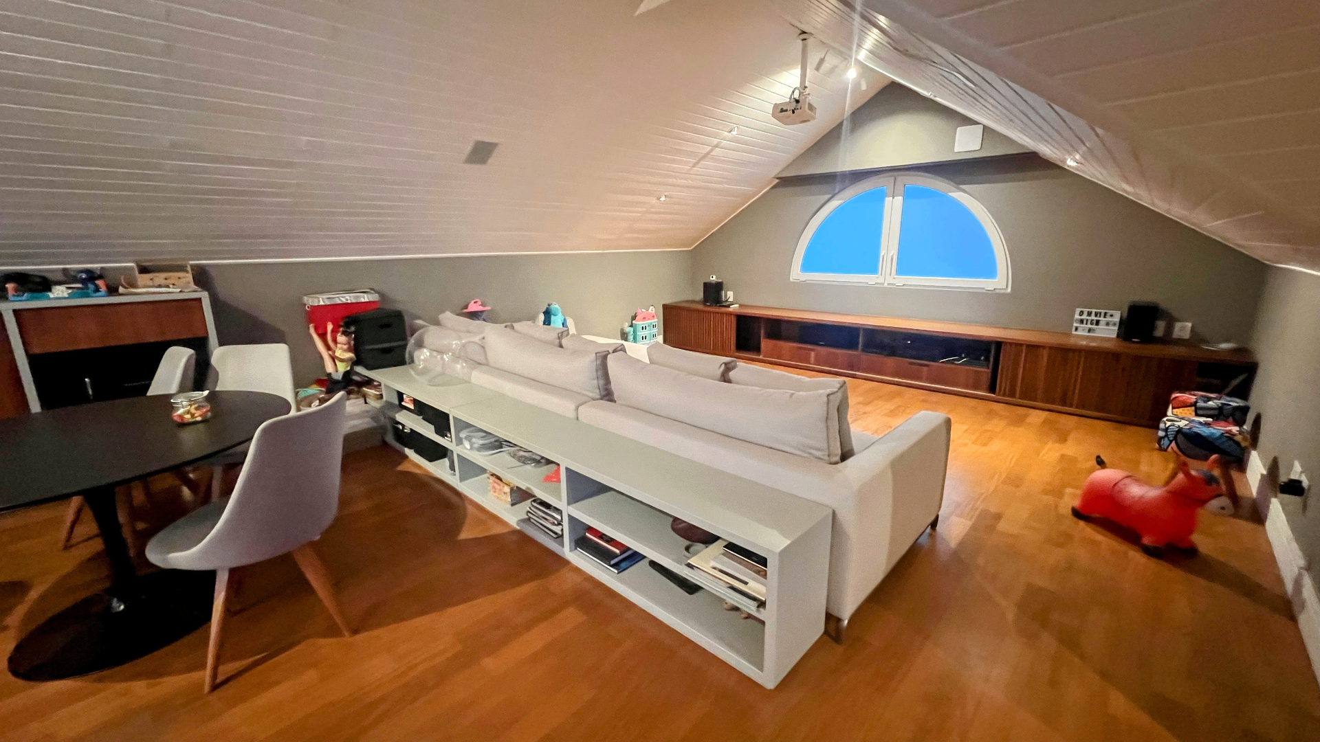 Sótão configurado como lounge com sofá cinza, projetor e janela em arco